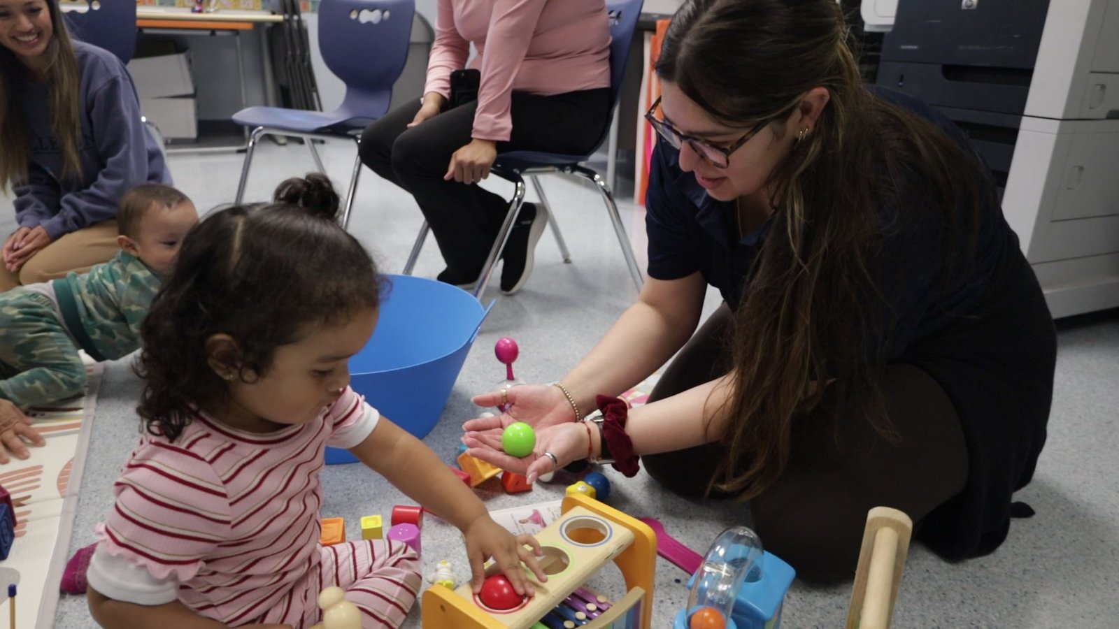 A lab member kneeling with a toddler during a hands-on assessment activity, using a wooden ball-drop toy